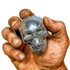 Echter Pyrit-Totenkopf in kräftiger, schmutzverschmierter Männerhand – polierter Wikinger-Schädel aus Stein, Symbol für Mut, Stärke, Schutz und nordische Handwerkskunst.