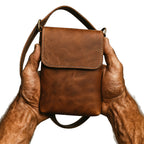 Muskulöse, adrig-männliche Wikingerhände halten eine handgefertigte Umhängetasche aus echtem Rindsleder – Modell ODIN in Braun, Nordskulls Stil