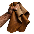 Kräftige, muskulöse Männerhand hält ein Stück naturbelassenes, braunes Echtleder vor reinweißem Hintergrund. Detailaufnahme mit sichtbaren Adern und Struktur – Symbol für nordische Handwerkskraft im NORDSKULLS-Stil.